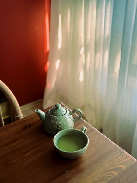 Grüne Teekanne und Tasse auf einem Holztisch neben einem sonnigen Fenster.